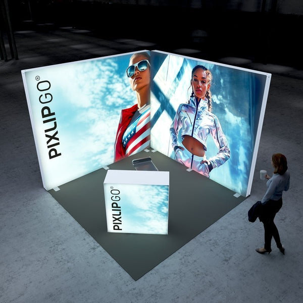 A bird's-eye view of the Pixlip GO exhibition booth. The setup includes two illuminated walls with vibrant images of stylish individuals against a sky backdrop, emphasizing energy and confidence. The booth features a small counter with minimalistic branding. A person is walking nearby.
