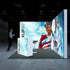 A dynamic exhibition booth by Pixlip GO featuring two large illuminated walls with bold imagery. The left wall showcases a confident woman in sunglasses and a patriotic outfit against a blue sky backdrop, while the right wall displays a sporty woman in a reflective tracksuit. A person approaches the booth holding a coffee cup.