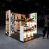 A modern exhibition booth by Pixlip GO, illuminated with large lightbox walls. The booth showcases premium coffee products, such as jars of coffee beans and packaging. In the foreground, there is a lit counter featuring product images. The background displays a cozy, rustic design with wooden shelves and decor.