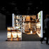 A frontal view of the Pixlip GO exhibition booth. The illuminated front of the counter displays coffee beans in jars and bags. The back walls are adorned with large graphics of a cozy café setting. A person carrying a bag is observing the booth from the side.