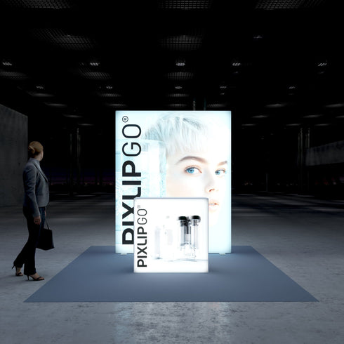 A frontal view of the Pixlip GO booth highlighting its illuminated backdrop with a striking portrait of a person with light hair and vibrant blue eyes. The compact counter in the foreground displays transparent bottle designs. A person walks past the booth carrying a bag.