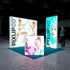 A frontal view of the Pixlip GO booth, emphasizing its playful and bright theme. The large illuminated wall features two women in an energetic foam battle, while the side panel highlights a portrait of a woman with a clean and fresh aesthetic. The counter displays a cheerful woman with a toothbrush in hand. A person stands nearby.
