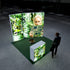 A Pixlip GO exhibition booth with a green, eco-friendly theme. The main illuminated panel features a close-up of a woman holding a circuit board in a lush forest setting. A side panel displays detailed greenery and technology. The front counter features similar visuals of circuit boards intertwined with nature. A person observes the setup from a distance.