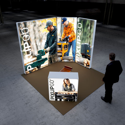 A Pixlip GO exhibition booth showcasing woodworking themes. The large illuminated panels feature images of two individuals operating wood-cutting and splitting machinery in outdoor settings. The front counter displays a woman working with wood on a table saw. A person stands nearby, observing the booth.