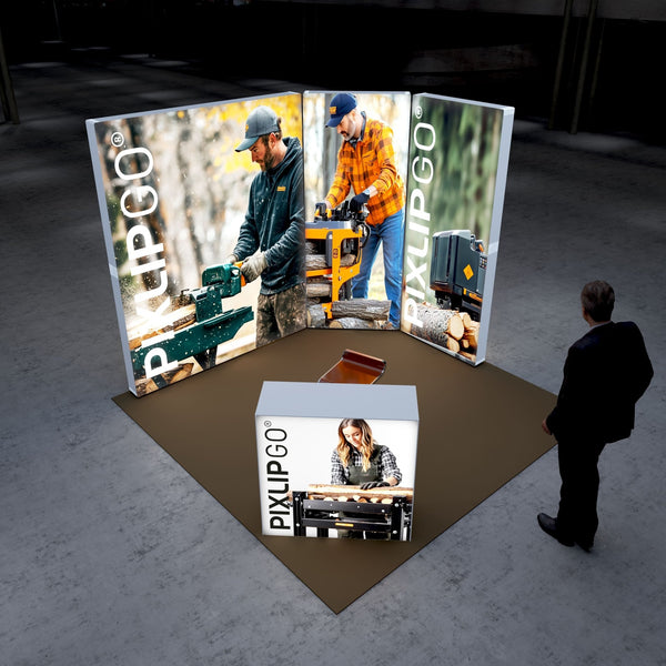 A Pixlip GO exhibition booth showcasing woodworking themes. The large illuminated panels feature images of two individuals operating wood-cutting and splitting machinery in outdoor settings. The front counter displays a woman working with wood on a table saw. A person stands nearby, observing the booth.