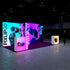 A side perspective of the exhibition setup, presenting a sleek design with glowing displays featuring high-tech camera imagery. The layout includes a magenta floor, lit display cubes, and a sophisticated environment emphasizing cutting-edge technology.