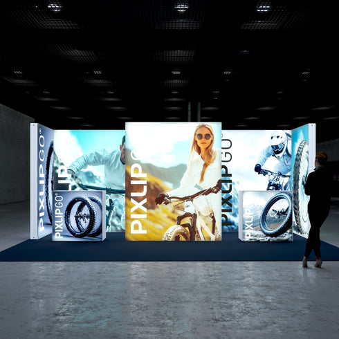 Front-facing view of a large illuminated booth, focusing on outdoor cycling enthusiasts. The center features a smiling woman with a bike against a scenic mountain backdrop. The sides display a male cyclist and detailed images of bike tires, emphasizing adventure and durability.
