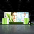 A frontal view of the Pixlip GO booth showcasing its focus on natural and scientific elements. The central illuminated backdrop combines imagery of natural products and laboratory expertise, while the counters complement the visuals with bright green accents. A person walks near the booth.