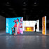 Front view of an exhibition stand with colorful illuminated displays. Models with trendy sunglasses and stylish outfits dominate the central blue panels, while a glowing orange display highlights a sleek handbag. The overall design emphasizes modern fashion and bright aesthetics.