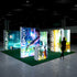 A side-angle view of the trade show setup, highlighting the illuminated lightboxes with detailed natural scenery and a glowing yellow accent panel. A woman is engaged with the booth, reflecting an interest in the environmental storytelling conveyed by the display