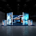 A straight-on view of the trade show booth with large illuminated panels showcasing a futuristic aircraft, robotic machinery in an industrial setting, and advanced technology interfaces. The booth is well-lit with a dark background for contrast, creating a professional and cutting-edge atmosphere.