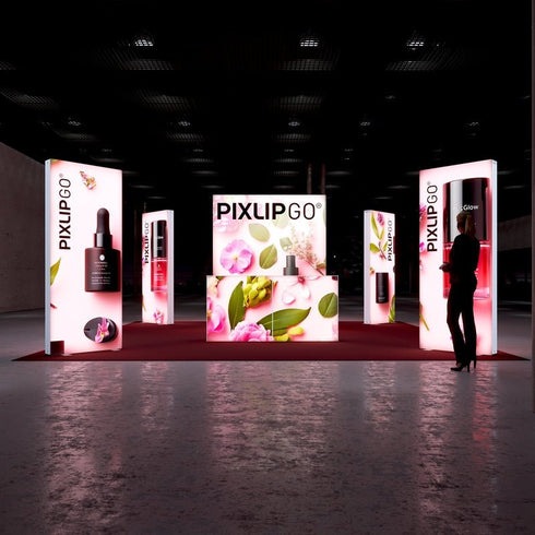 A frontal view of the exhibition booth with vibrant pink flower graphics, showcasing cosmetic products such as droppers and bottles. The PIXLIP GO branding is prominent on all lit panels, while the floor is a deep red carpet.