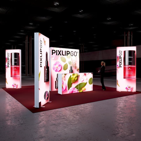 A brightly illuminated exhibition booth featuring a floral and beauty product theme. The main panel showcases a variety of pink flowers and cosmetic bottles, framed with glowing panels and the PIXLIP GO branding. A person in professional attire observes the booth.